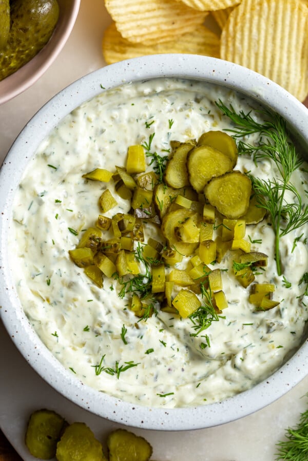Dill pickle dip in a bowl topped with bright green diced pickles. There are ruffles chips in the corner of the shot