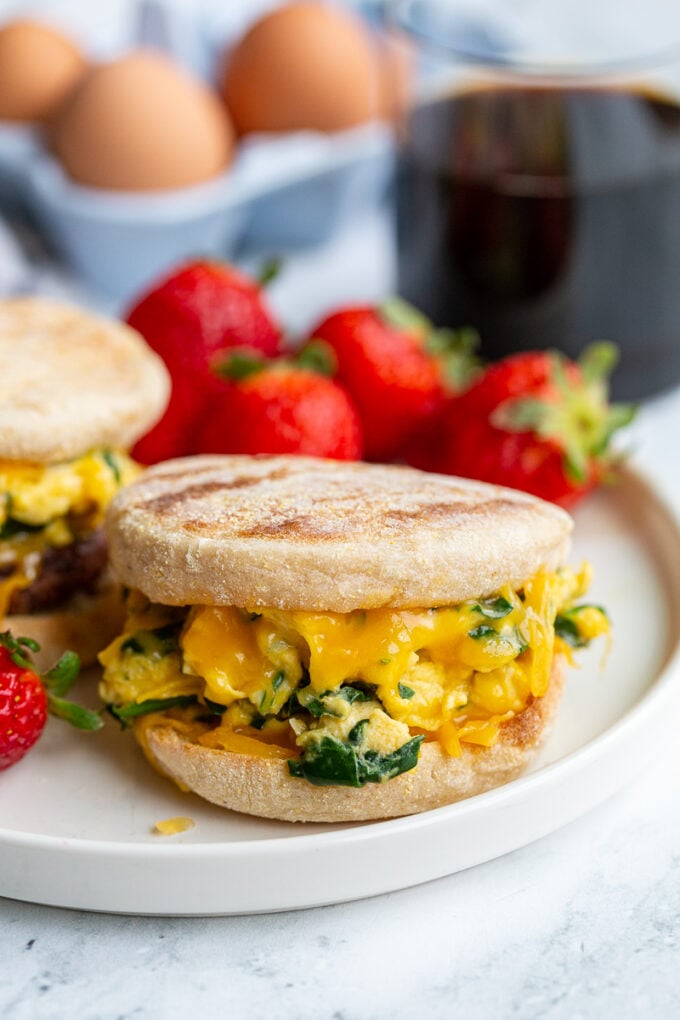 english muffin sandwich with spinach scrambled eggs topped with cheese in the middle. strawberries and coffee are in the background 