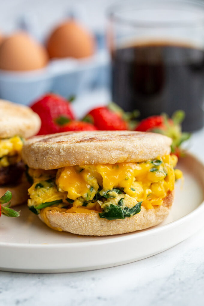 english muffin sandwich with spinach scrambled eggs topped with cheese in the middle. strawberries and coffee are in the background